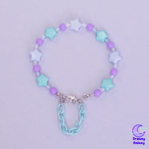 Könnte beinhalten: Ein Armband mit Stern- und runden Perlen in Pastellblau, Lila und Weiß. Das Armband hat einen silberfarbenen Verschluss und einen hellblauen Kettenanhänger. Das Dreamy Galaxy-Logo befindet sich unten rechts.