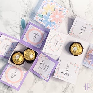 May include: A handmade, open gift box with a purple frame, containing two gold-wrapped chocolates. The box is decorated with floral designs, pastel colors, and Italian text. The box is designed to be given as a gift.