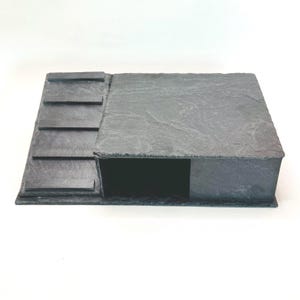 May include: A dark grey slate structure featuring a flat, textured square platform attached to a ramp section. The ramp includes four horizontal raised steps leading up to the main elevated surface, which has a hollow opening underneath the top slab.