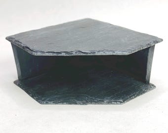 May include: A dark gray slate corner shelf featuring a triangular top surface with a rough, natural stone texture and jagged edges. The structure includes a matching base plate connected by two side panels, creating an open, hollow interior space.
