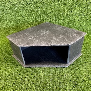 May include: A dark grey corner shelf with a textured, stone-like finish. The shelf features a triangular top surface and an open, hollow front section, designed to fit into a corner space with a flat, wide opening for storage or display purposes.