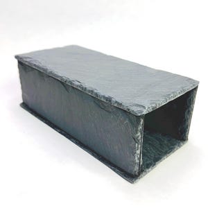 May include: A rectangular tunnel-shaped structure constructed from dark grey slate stone. The object features a rough, natural textured surface on the exterior and top, with an open hollow centre running through the length of the piece.