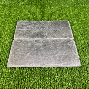 May include: Two pieces of dark grey slate tile, stacked on top of each other, with a rough, textured surface. The tiles are on a green artificial grass background.
