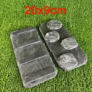 May include: Two rectangular, dark grey slate serving platters, each measuring 20 x 9 cm. One has three raised horizontal bars, the other has four decorative stone accents. The text "20 x 9cm" is in red.