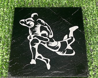 May include: Black and white silhouette of two rugby players in action on a black slate coaster.