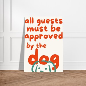 Könnte beinhalten: Ein rechteckiges Poster mit dem Text "all guests must be approved by the dog" in Rot, mit einer Cartoon-Hundeillustration in Türkis und Weiß. Das Poster wird vor einer weißen Wand und einem Holzboden angezeigt.