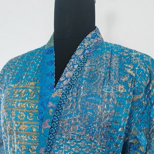 Op de afbeelding: Een blauwe badjas met patroon, bijpassende riem en zakken. De badjas wordt getoond op een zwarte mannequin. De stof heeft een patchwork ontwerp met gouden en witte accenten. De badjas is gemaakt van een zacht, lichtgewicht materiaal.