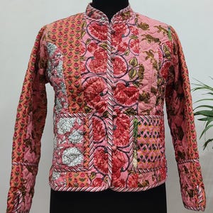 May include: A pink quilted jacket with a floral design in red and green. The jacket features a mandarin collar and long sleeves. The floral pattern is consistent across the jacket, creating a cohesive design.