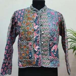 Puede incluir: Chaqueta acolchada con estampado floral en tonos azules, rosas y naranjas. La chaqueta tiene cuello mandarín, mangas largas y cierre de botones. La chaqueta se exhibe en un maniquí negro.