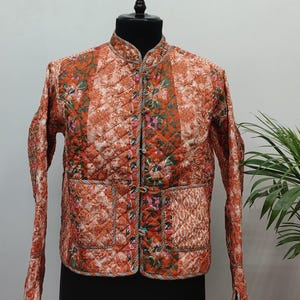 Puede incluir: Chaqueta acolchada en tonos naranja, rojo y verde, con un estampado floral. La chaqueta tiene cuello mandarín, mangas largas y dos bolsillos delanteros. Se exhibe en un maniquí negro sobre un fondo neutro.