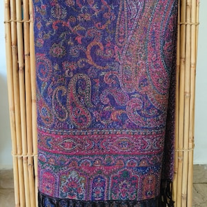 Puede incluir: Bufanda con estampado de cachemir en tonos morado, fucsia y turquesa. La bufanda presenta un diseño intrincado con un borde con flecos. El textil está drapeado sobre una estructura de bambú, mostrando sus ricos colores y su patrón detallado.