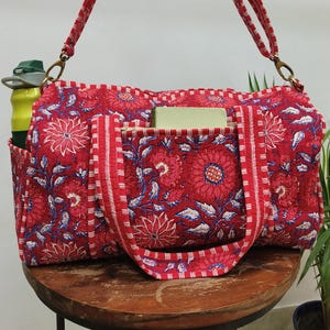 Puede incluir: Bolsa de deporte roja con estampado floral y correa a juego. La bolsa tiene un bolsillo lateral que contiene una botella de agua amarilla. La bolsa tiene forma rectangular con un estampado floral en rojo y azul. La bolsa es de tela acolchada.