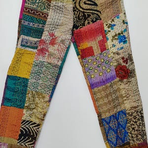 Pode incluir: Calças patchwork com cintura com cordão. As calças são feitas de vários retalhos de tecido em uma variedade de cores, incluindo rosa, azul, amarelo e laranja. Os retalhos têm diferentes padrões e texturas. As calças estão penduradas em um cabide de madeira.