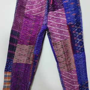 May include: Purple patchwork pants with a drawstring waist. The pants feature a variety of purple, blue, and pink fabric patches, stitched together with white thread. The pants are hanging on a wooden hanger against a white background.