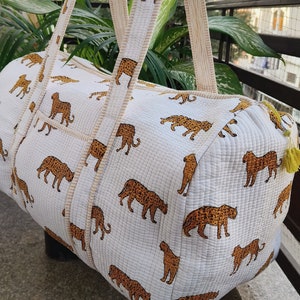 May include: White quilted duffel bag with brown leopard print. The bag has two long straps with the same print. The bag is hanging on a wooden hanger.