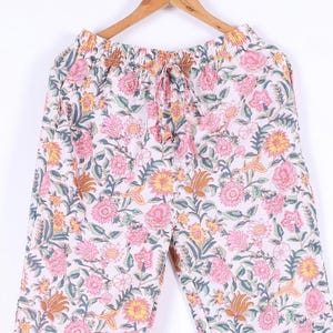 May include: White pajama pants with a floral pattern in pink, yellow, and green. The pants are hanging on a wooden hanger. The floral design covers the entire surface of the pants.
