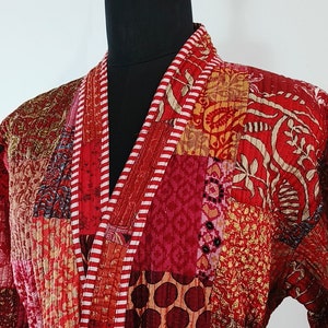 Puede incluir: Una bata roja acolchada con un diseño de patchwork. La bata tiene un cinturón de corbata a rayas blancas y rojas y dos bolsillos.