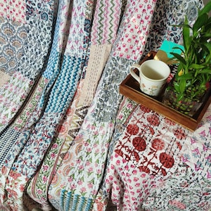 May include: A white kantha quilt with a patchwork design featuring various floral and geometric patterns in shades of blue, pink, red, and green. The quilt is draped over a chair and a wooden tray with a cup and a potted plant.