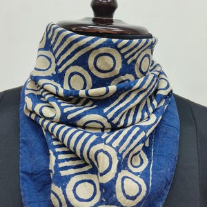 May include: A blue and cream scarf with a geometric pattern of circles and stripes. The scarf is draped around a wooden mannequin head. The scarf's border is a solid blue colour.