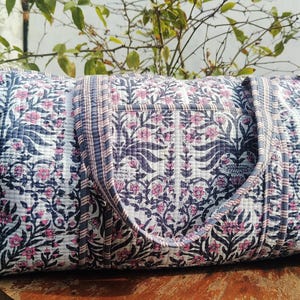 May include: A quilted travel bag with a floral pattern in navy blue, white, and pink. The bag has a rectangular shape with a top handle and a shoulder strap. The bag is made of a soft, quilted fabric.