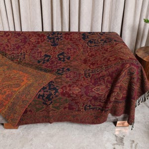 Könnte beinhalten: Eine rechteckige Wohndecke mit einem komplexen Paisley-Muster in den Farben Dunkelrot, Bordeaux, Marineblau und Dunkelgrün. Eine Ecke ist umgeschlagen und zeigt ein kontrastierendes orange-goldenes Paisley-Design mit Fransenbesatz.