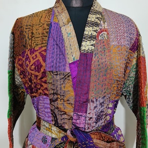 May include: A patchwork robe in a variety of colors, including purple, gold, and green. The robe is made of different fabric squares sewn together. It has long sleeves and a matching belt. The robe is displayed on a black mannequin.
