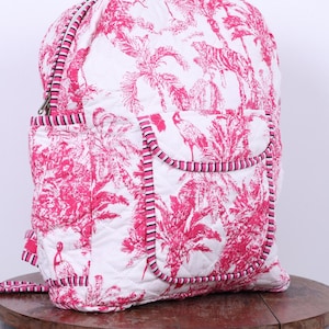 Puede incluir: Mochila acolchada blanca y rosa con estampado tropical de palmeras, pájaros y tigres. La mochila tiene un bolsillo frontal y está ribeteada con ribetes a rayas rosas y negras. La bolsa está sobre una mesa de madera.