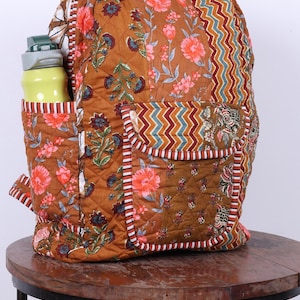 May include: A floral-patterned quilted backpack in brown, pink, and blue. It features a side pocket with a yellow water bottle and a front pocket with a zigzag design. The bag has white and red trim and is suitable for everyday use.