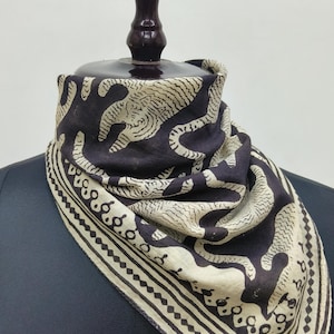 Könnte beinhalten: Ein dunkelbrauner und cremefarbener quadratischer Schal mit einem sich wiederholenden Muster stilisierter Tierfiguren. Der Schal ist über einer Schaufensterpuppe drapiert und zeigt sein Design und seine Textur. Die Kanten haben einen dekorativen Rand.