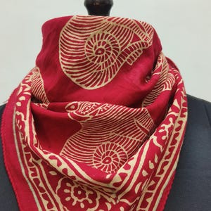 May include: A red scarf with a beige pattern. The scarf displays a repeating design of beige spiral and floral patterns on a red background. The scarf is draped over a black mannequin.