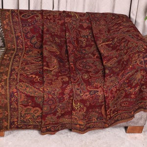 May include: A rectangular throw blanket featuring a deep red base colour with an intricate, multi-coloured paisley pattern in shades of gold, orange, and dark brown. The edges of the blanket are finished with dark brown fringe along the short sides.