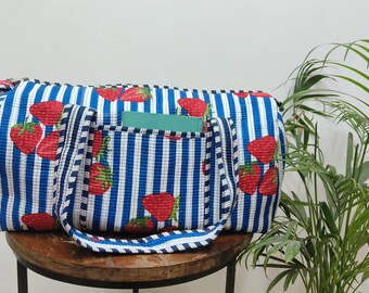 Puede incluir: Bolso tote a rayas azules y blancas con estampado de fresas. El bolso tiene un bolsillo delantero y asas. Un cuaderno verde es visible en el bolsillo. El bolso está sobre un taburete de madera.