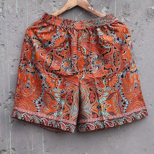 May include: Orange, black, and teal shorts featuring a dense, intricate paisley and floral pattern throughout. The shorts have an elasticated waistband and a wide, decorative border along the hemline, hanging from a wooden clothes hanger.