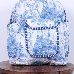 Könnte beinhalten: Ein gesteppter Rucksack mit blau-weißem Toile-de-Jouy-Muster, das eine Dschungelszene darstellt. Der Rucksack hat eine Klappentasche und zwei Seitentaschen, alle mit blau-weiß gestreiftem Stoff eingefasst. Er steht auf einem runden Holztisch.