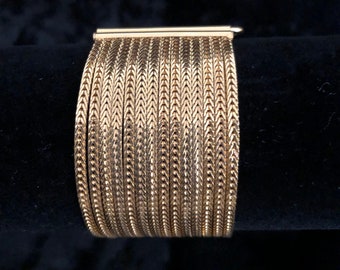 May include: A gold-toned metal bracelet with a woven design. The bracelet is made of multiple thin strips of metal that are woven together to create a herringbone pattern.
