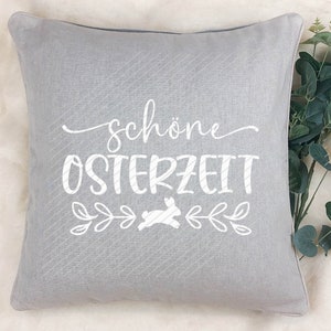 Op de afbeelding: Een grijs kussen met de tekst "sch&ouml;ne Osterzeit" en een witte konijnenafbeelding. Het kussen heeft een subtiel gestreept patroon.