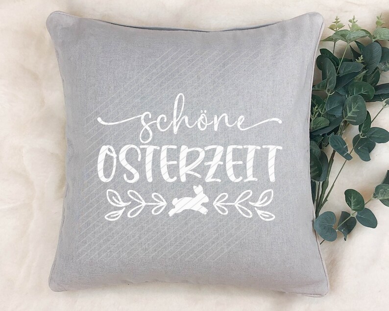 Op de afbeelding: Een grijs kussen met de tekst "sch&ouml;ne Osterzeit" en een witte konijnenafbeelding. Het kussen heeft een subtiel gestreept patroon.