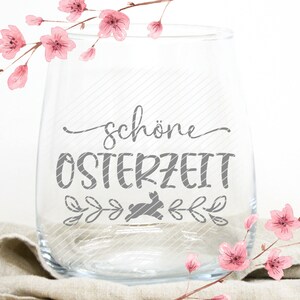 Op de afbeelding: Een helder glazen wijnglas met de tekst "sch&ouml;ne Osterzeit" in grijze letters. Onder de tekst bevindt zich een grijs silhouet van een konijn. Het glas is omringd door roze aquarelbloemen.