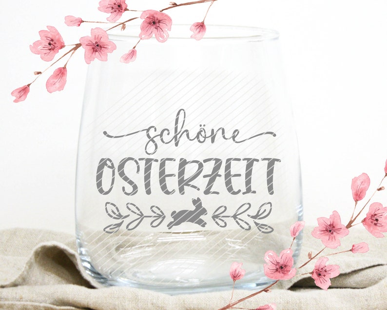 Op de afbeelding: Een helder glazen wijnglas met de tekst "sch&ouml;ne Osterzeit" in grijze letters. Onder de tekst bevindt zich een grijs silhouet van een konijn. Het glas is omringd door roze aquarelbloemen.