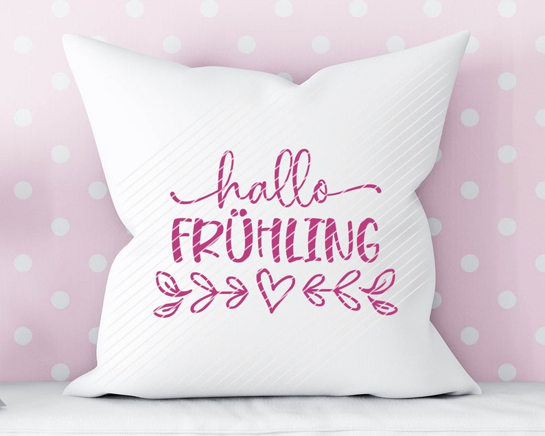 K&ouml;nnte beinhalten: Wei&szlig;es Kissen mit rosa Schriftzug "hallo FR&Uuml;HLING" mit einem rosa Herz und einem Blattdesign.