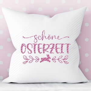 Op de afbeelding: Een wit kussen met roze tekst die "sch&ouml;ne Osterzeit" zegt met een roze konijnen silhouet en bloemmotieven.