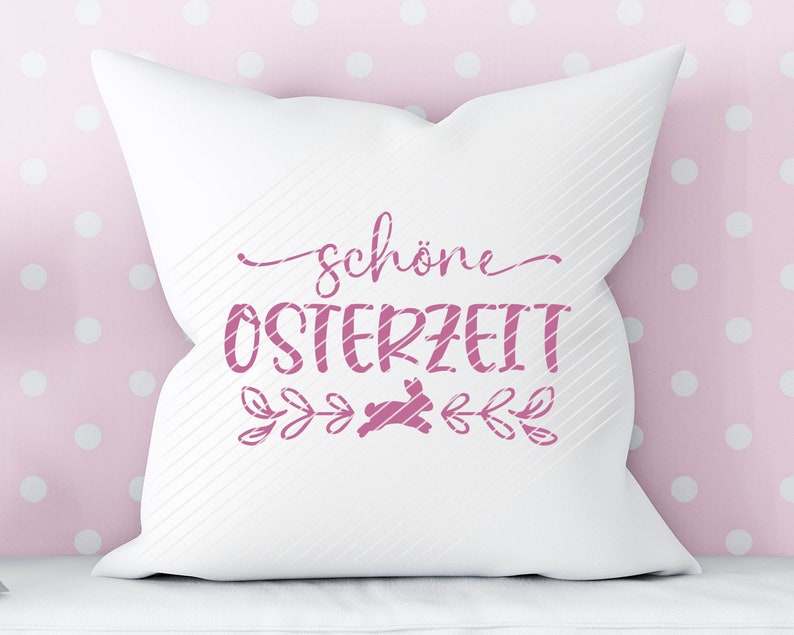 Op de afbeelding: Een wit kussen met roze tekst die "sch&ouml;ne Osterzeit" zegt met een roze konijnen silhouet en bloemmotieven.
