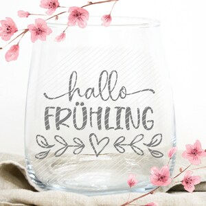 K&ouml;nnte beinhalten: Ein klares Weinglas aus Glas mit dem Text "hallo FR&Uuml;HLING" in grauer Schrift. Das Glas ist mit kleinen grauen Bl&auml;ttern und einem Herzen verziert. Das Glas steht auf einer beigefarbenen Stoffoberfl&auml;che. Rosa Aquarellblumen sind im Hintergrund.
