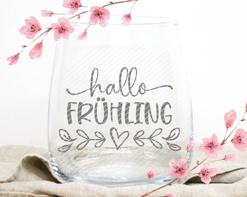 K&ouml;nnte beinhalten: Ein klares Weinglas aus Glas mit dem Text "hallo FR&Uuml;HLING" in grauer Schrift. Das Glas ist mit kleinen grauen Bl&auml;ttern und einem Herzen verziert. Das Glas steht auf einer beigefarbenen Stoffoberfl&auml;che. Rosa Aquarellblumen sind im Hintergrund.