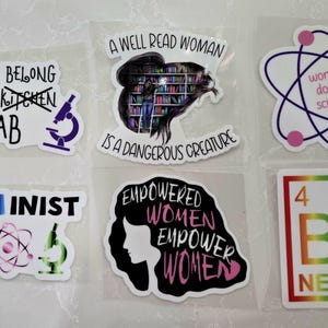 Könnte beinhalten: Eine Sammlung von Aufklebern mit ermächtigenden Botschaften und Designs zum Thema Wissenschaft. Die Aufkleber zeigen Sätze wie "Women Belong in the Lab", "STEMinist" und "Be Nerdy". Weitere Designs sind eine Frau in einer Bibliothek und ein Atomsymbol.