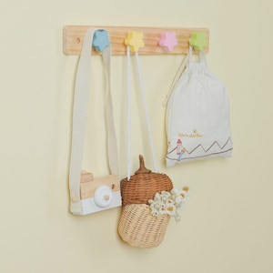 Puede incluir: Un estante de pared de madera con cinco ganchos en forma de estrella en colores pastel. Una bolsa de lona blanca con el texto "wonderbee" cuelga de un gancho. Una pequeña cámara de madera con una lente blanca cuelga de otro gancho. Una cesta de mimbre con una tapa de forma de bellota marrón cuelga de un tercer gancho.