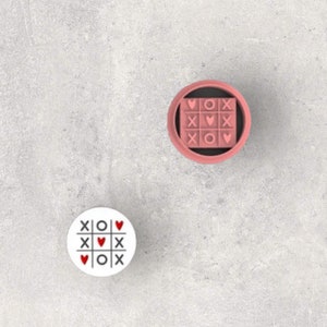 May include: Two white round game boards with a tic-tac-toe design. The game pieces are hearts and X's. One board is in a pink circle.