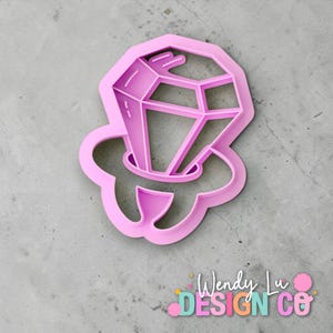 May include: A pink plastic cookie cutter in the shape of a diamond engagement ring. The cutter has a detailed design with facets on the diamond and a ring band. The cutter is on a gray surface.