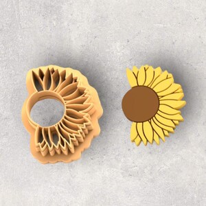 May include: Two sunflower-shaped cookie cutters. One is a light orange plastic cutter with a detailed sunflower design. The other is a yellow and brown sunflower-shaped decoration. The background is a light gray surface.
