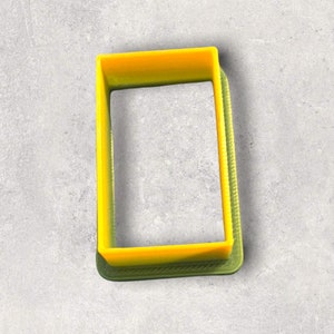 May include: A bright yellow rectangular cookie cutter with a textured edge. The cutter is made of plastic and is set against a gray background. The cutter is designed to cut out rectangular shapes from dough or fondant.